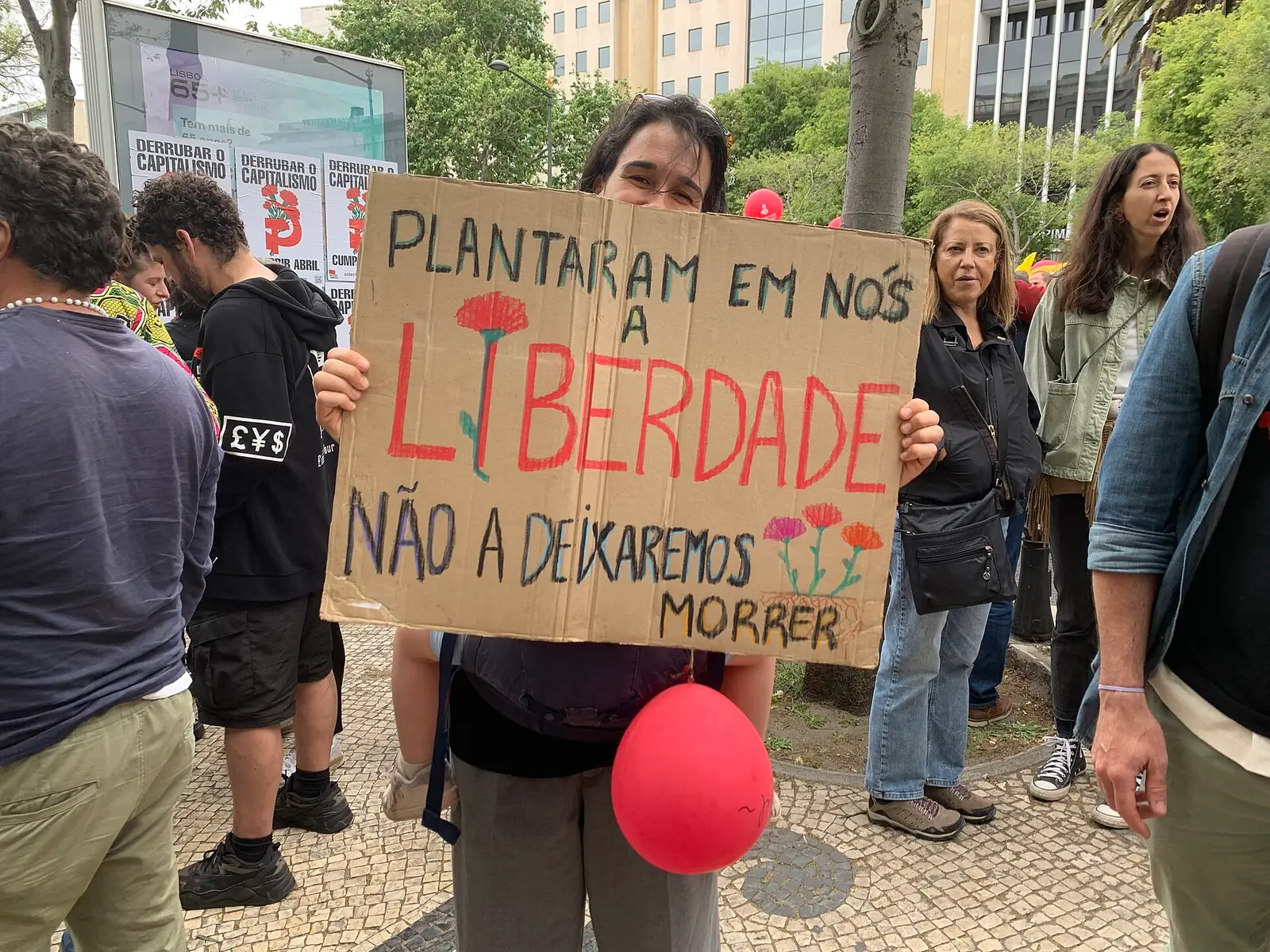 Uma pessoa segura à frente do peito um cartaz de cartão com a palavra «liberdade» a vermelho e cravos desenhados; à volta, gente na calçada e um balão vermelho.