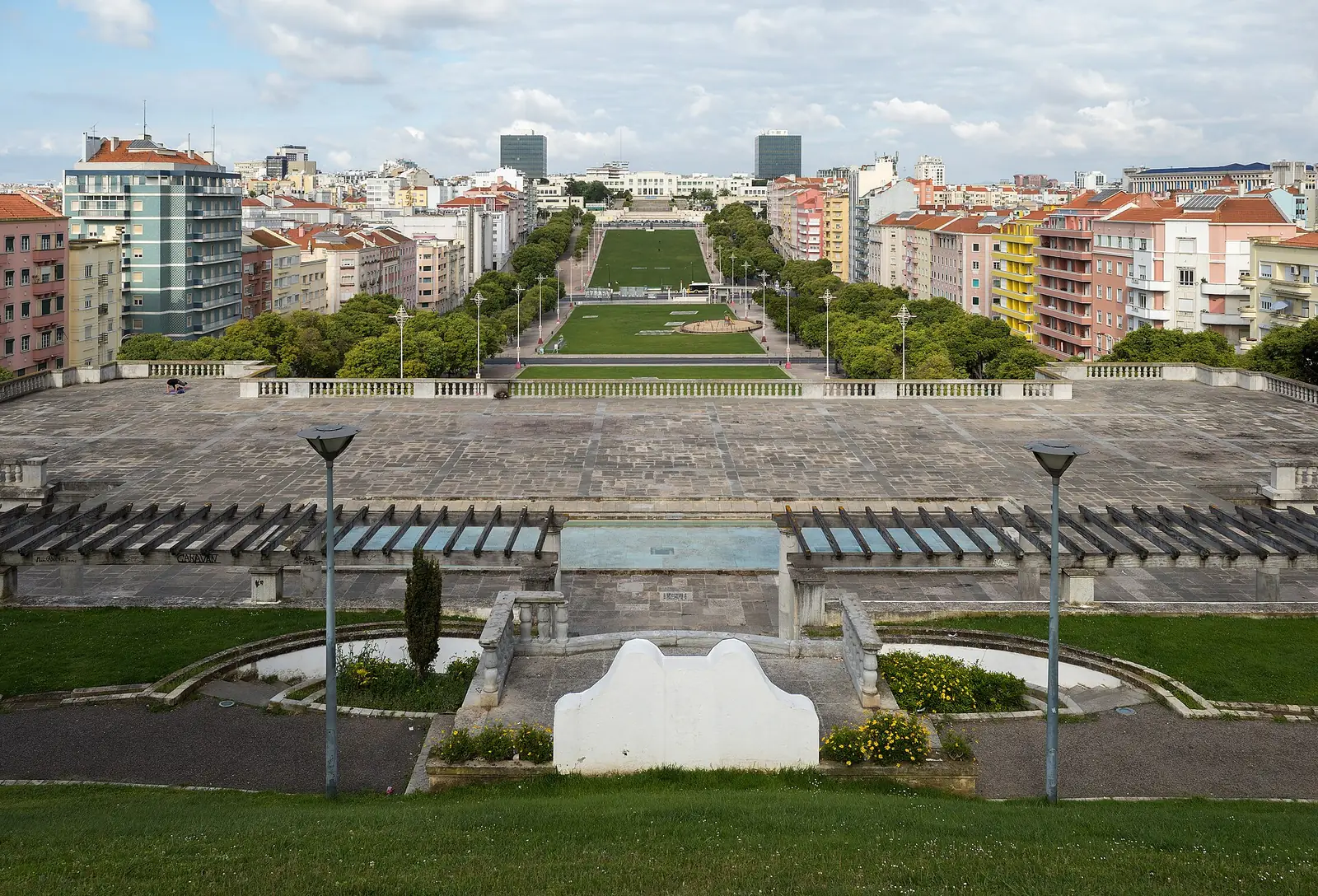 Vista do alto sobre um grande relvado comprido e vazio, entre avenidas e prédios, que se estende até edifícios brancos no extremo oposto.