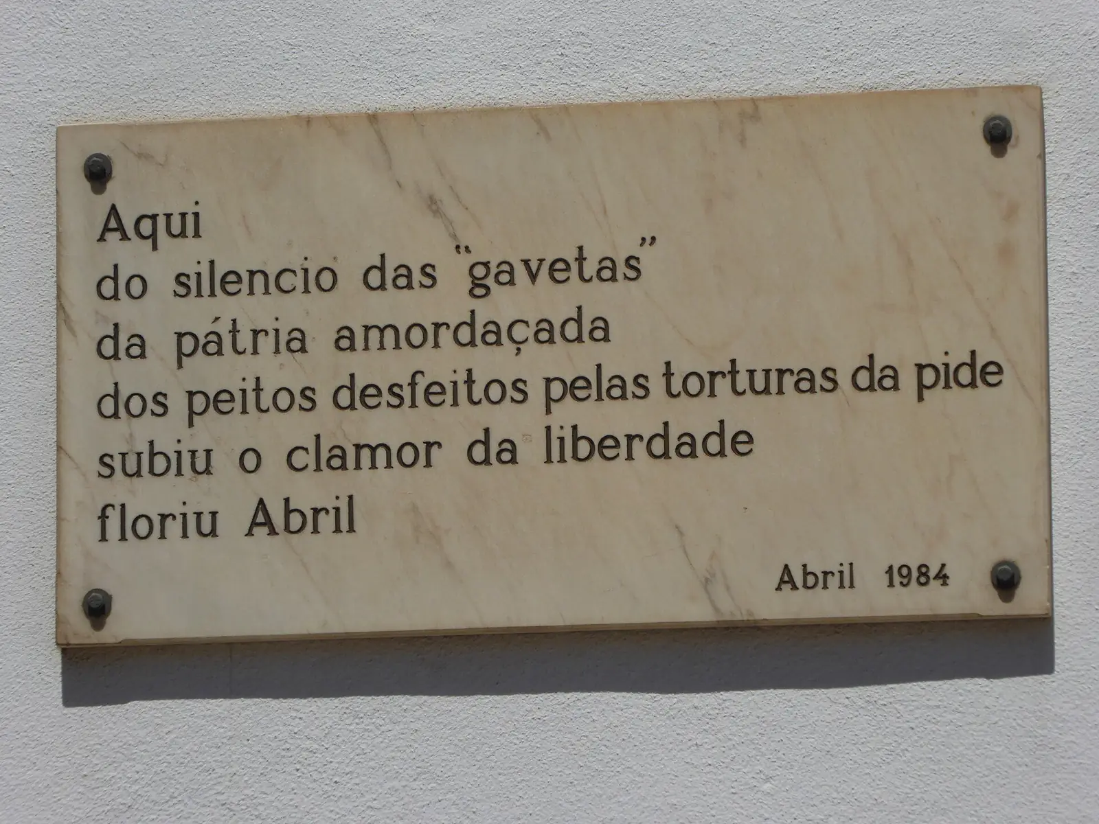 Placa de pedra clara presa a uma parede branca, com um poema curto gravado a negro sobre o silêncio das «gavetas», as torturas da PIDE e o Abril que floriu, assinado «Abril 1984».
