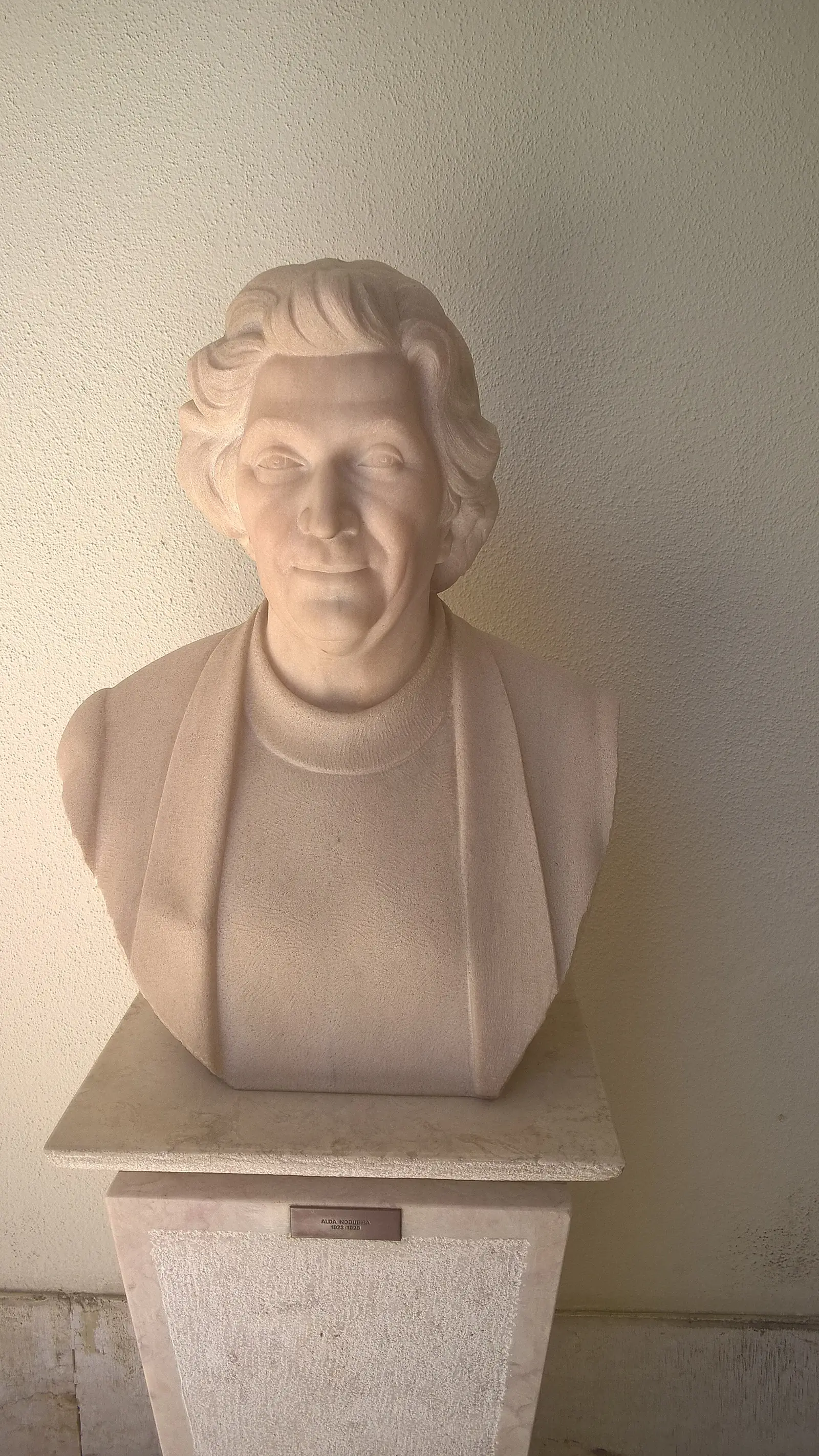 Busto de mármore branco de uma mulher de cabelo curto e ondulado, a sorrir, sobre um pedestal de pedra encostado a uma parede clara.