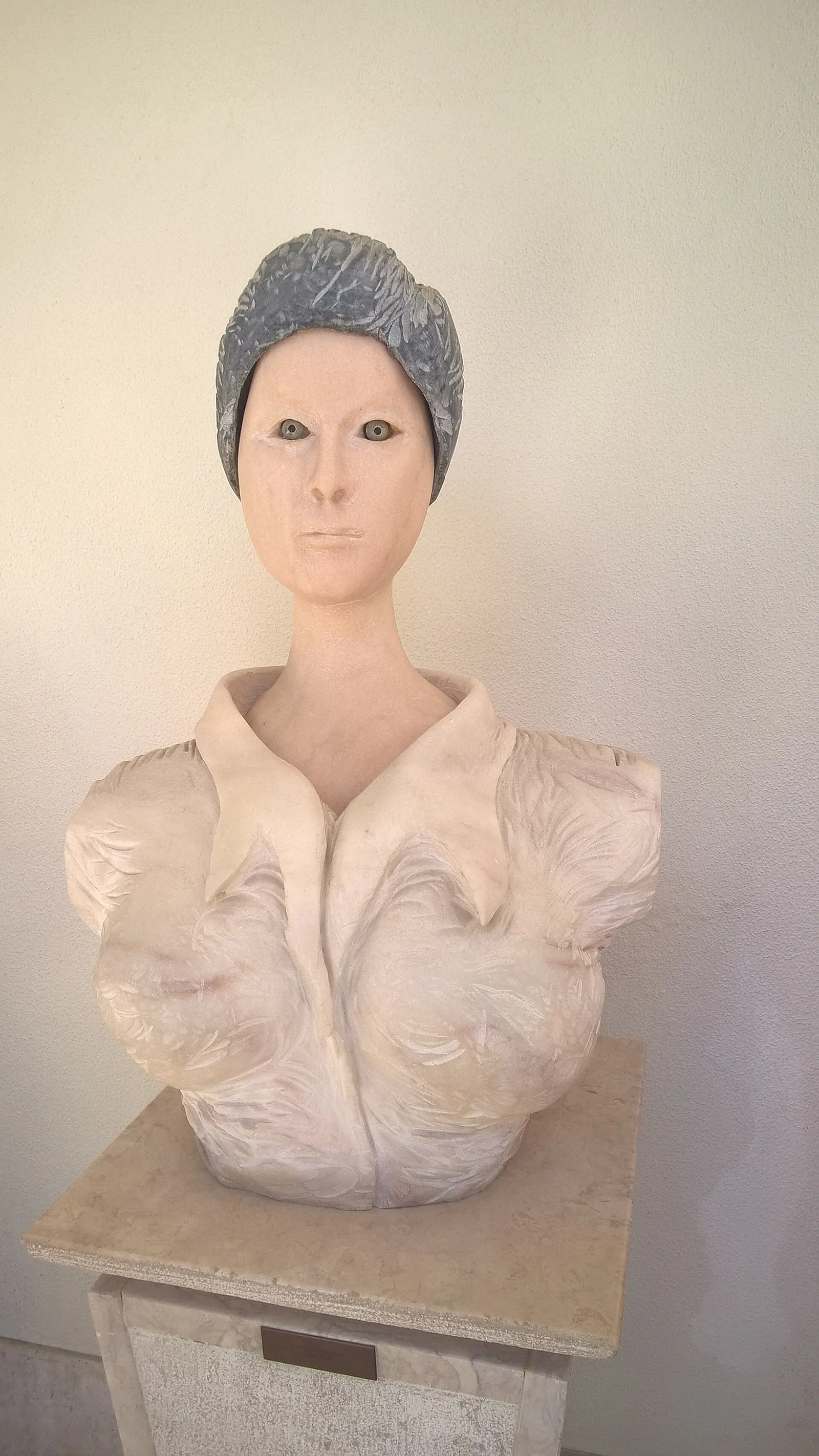 Busto de mármore estilizado de uma mulher de pescoço alto, olhos pintados e cabelo apanhado em pedra cinzenta, com uma gola de folhos esculpida, sobre um pedestal.