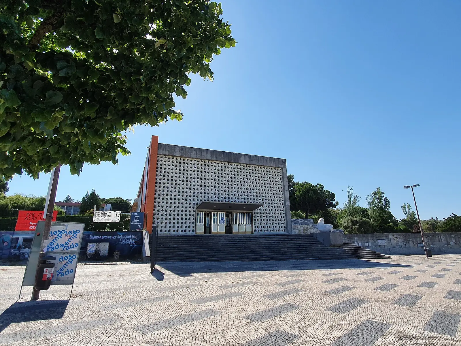 Fachada branca de blocos vazados com uma porta ao centro e uma ala de tijolo à esquerda, diante de um largo de calçada com árvores, sob céu azul.