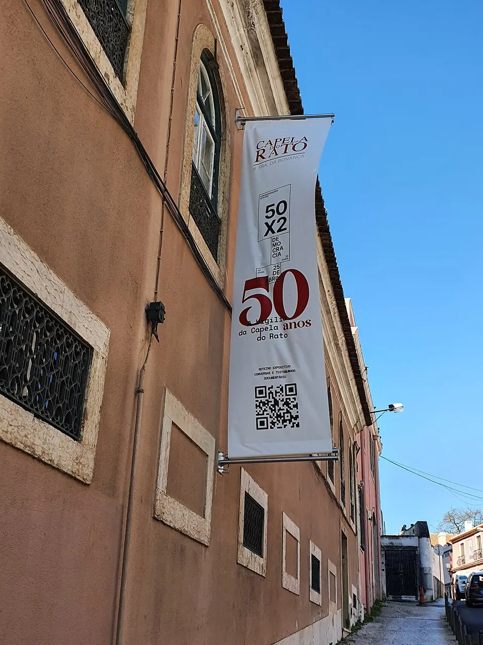 Bandeira vertical branca presa à fachada cor de salmão, com um grande 50 a vermelho e as palavras «Vigília da Capela do Rato»; ao fundo, a rua estreita a descer.