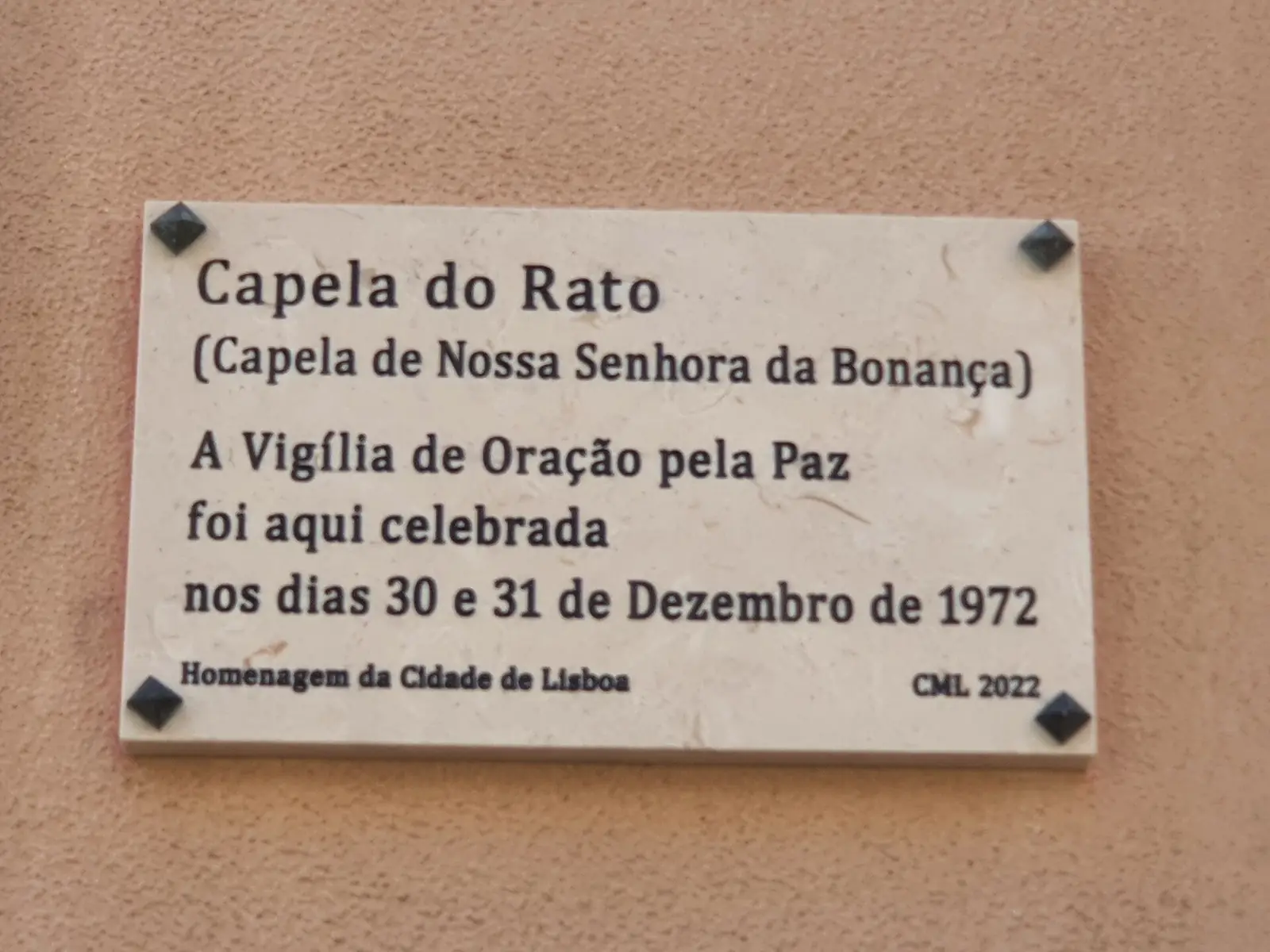 Placa de pedra clara numa parede cor de salmão, com o texto «Capela do Rato (Capela de Nossa Senhora da Bonança) – A Vigília de Oração pela Paz foi aqui celebrada nos dias 30 e 31 de Dezembro de 1972 – Homenagem da Cidade de Lisboa – CML 2022».