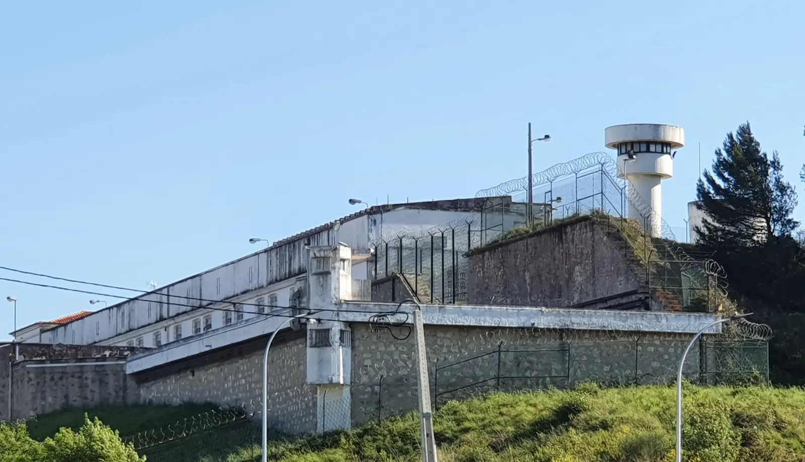 Muralha de pedra com uma vedação por cima, uma torre de vigia redonda e branca ao fundo e pinheiros, sob céu azul.