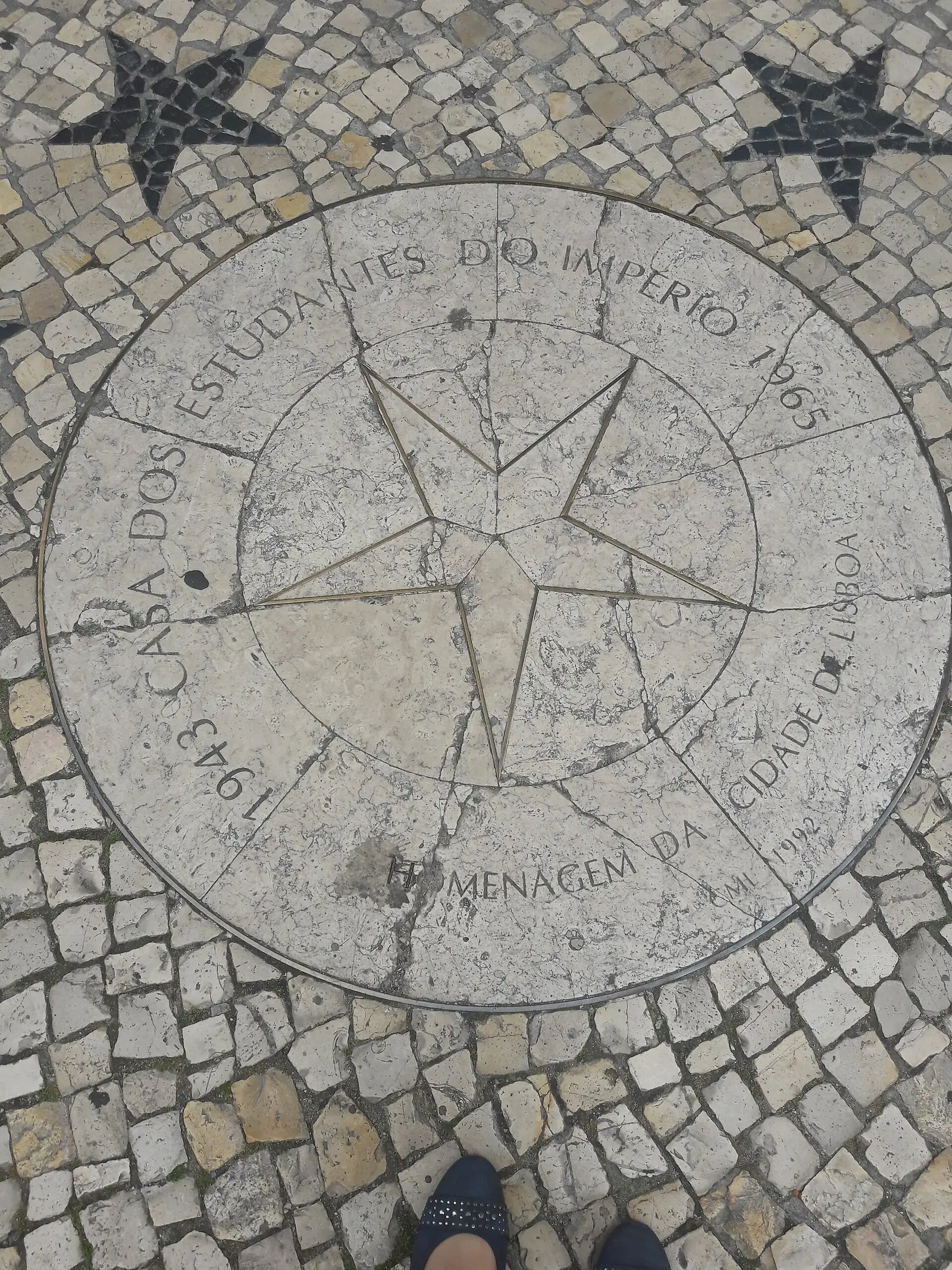 Disco de pedra clara embutido na calçada portuguesa, com uma rosa-dos-ventos gravada ao centro e a inscrição «Casa dos Estudantes do Império – 1943-1965 – Homenagem da Cidade de Lisboa – C.M.L. 1992» à volta.