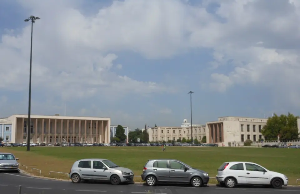 Edifícios compridos de pedra clara, de linhas direitas, do outro lado de um relvado e de uma fila de carros estacionados, sob céu com nuvens.