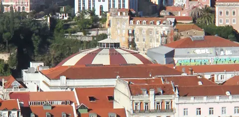 Vista do alto sobre telhados, com uma grande cúpula metálica arredondada entre prédios e árvores em primeiro plano.