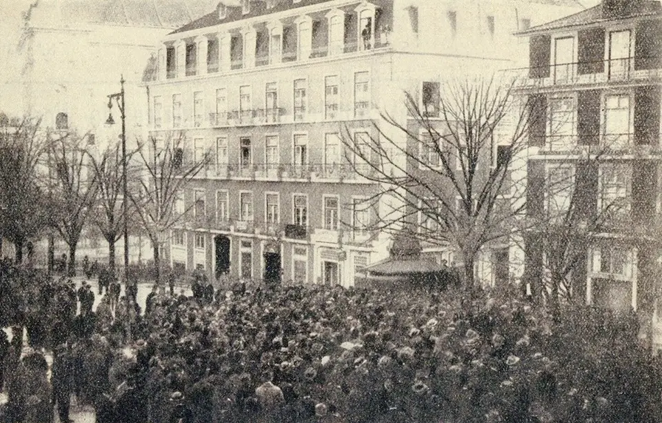 Fotografia antiga e amarelada de uma multidão compacta numa praça, diante de prédios altos e de árvores despidas.