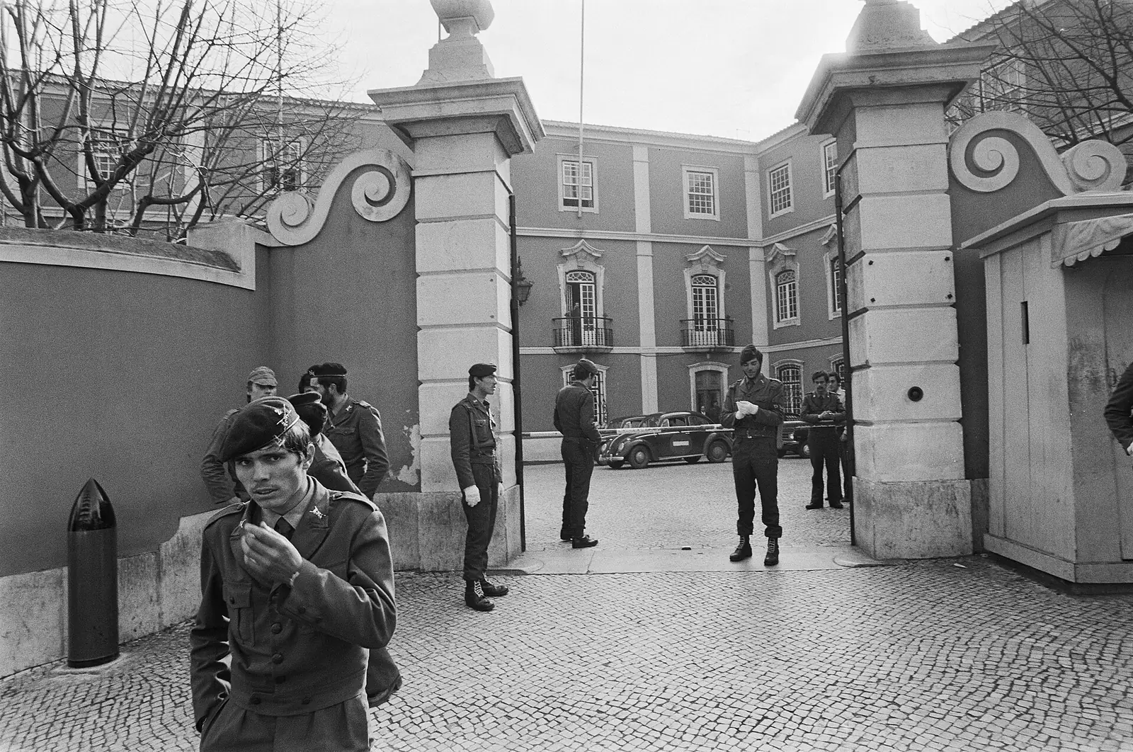 Fotografia a preto e branco de militares de capacete e farda junto a um portão de pilares de pedra, com o pátio e a fachada do palácio ao fundo.