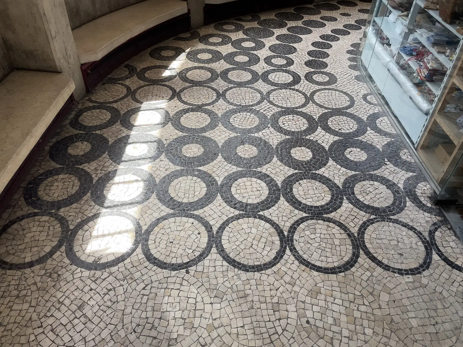 Chão de calçada portuguesa com um padrão de círculos negros sobre fundo branco, atravessado por uma faixa de sol, junto a uma montra.