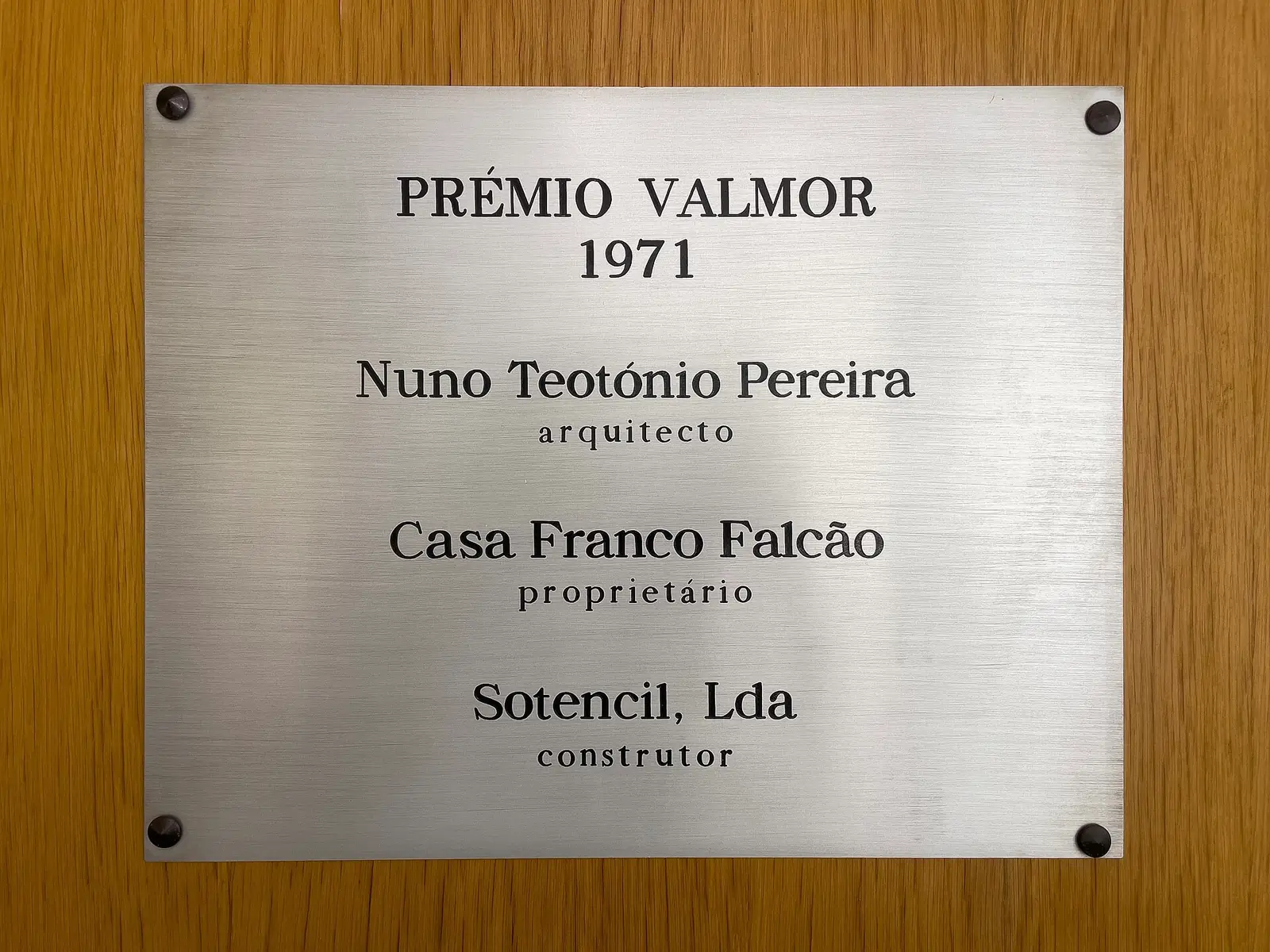 Placa metálica retangular presa a uma parede de madeira, com o texto gravado «Prémio Valmor 1971 – Nuno Teotónio Pereira, arquiteto – Casa Franco Falcão, proprietário – Sotencil, Lda, construtor».