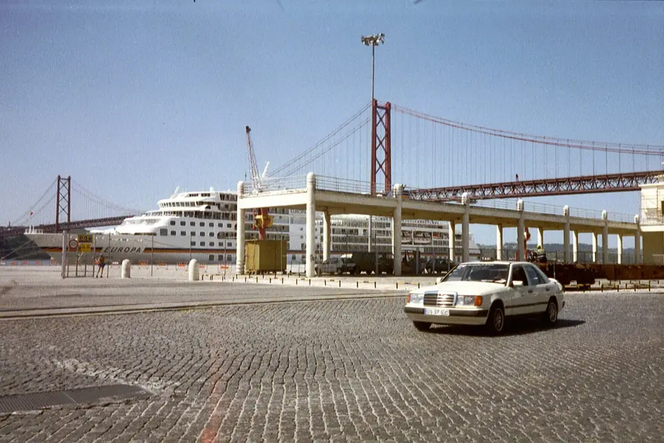 Um navio de cruzeiro branco atracado, por trás de uma passagem elevada de betão, sobre pilares, que corre ao longo do cais; por cima, a ponte suspensa vermelha; em primeiro plano, um largo empedrado com um automóvel branco.
