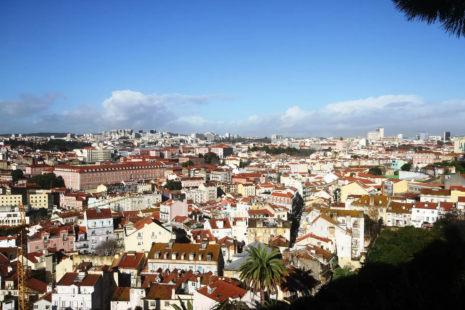 Vista do alto sobre telhados vermelhos, com um edifício comprido e rosado de muitas janelas ao centro e a cidade a estender-se até ao horizonte.
