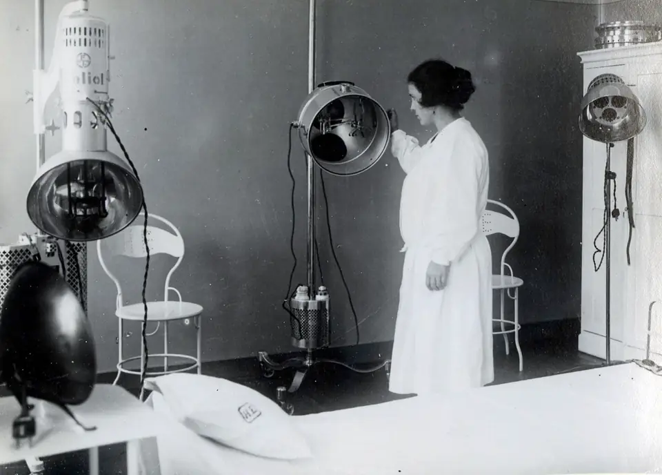 Fotografia a preto e branco de uma sala de hospital com grandes aparelhos de radiação suspensos e uma mulher de bata branca de pé junto a eles.