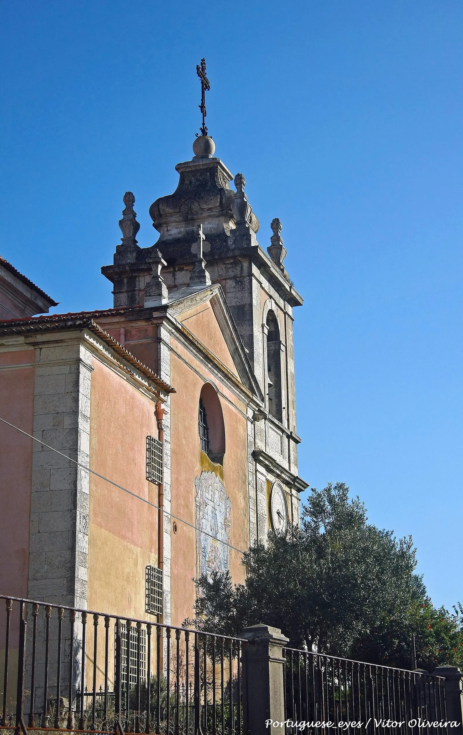 Torre sineira de pedra clara e fachada amarela de uma igreja, vistas de baixo contra um céu azul muito limpo, com uma árvore à direita.