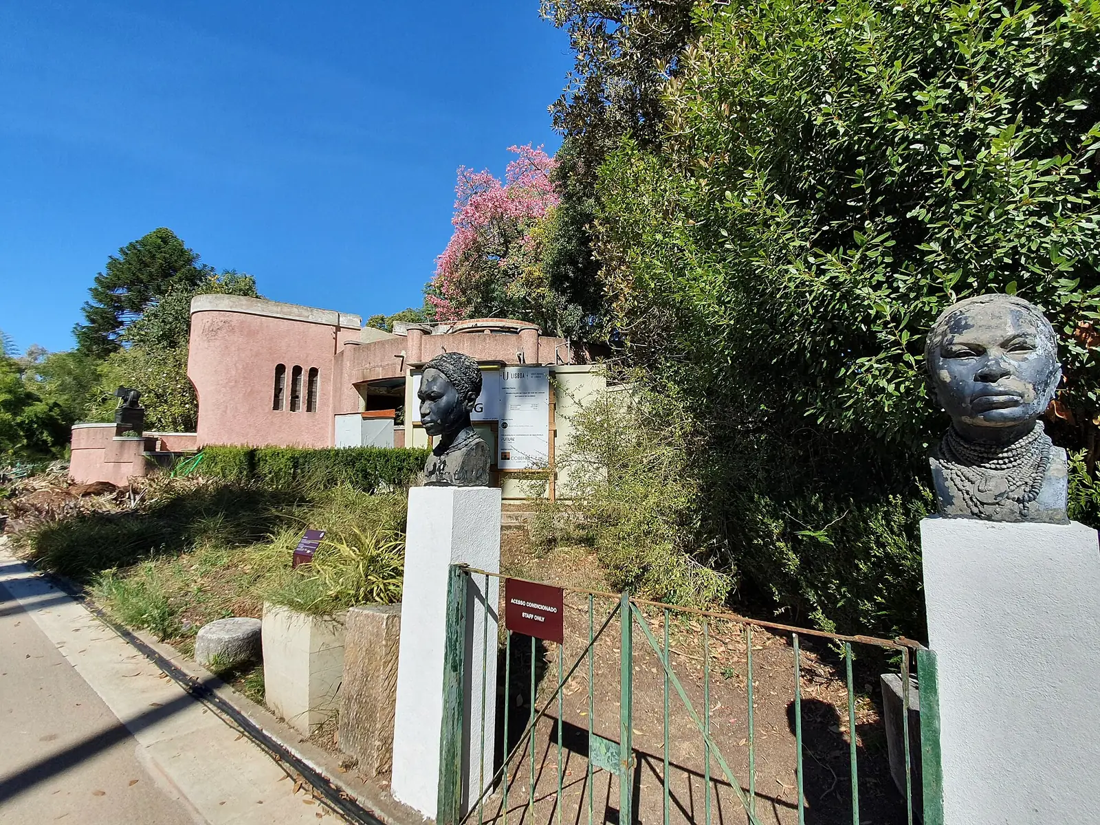Dois bustos escuros sobre pilares brancos, de cada lado de um portão de grades verdes: à direita, em primeiro plano, um rosto com muitos colares de contas ao pescoço; ao centro, outro busto, de cabelo entrançado. Ao fundo, entre arbustos e uma árvore de flores cor-de-rosa, um edifício cor-de-rosa de linhas curvas, com uma torre redonda.