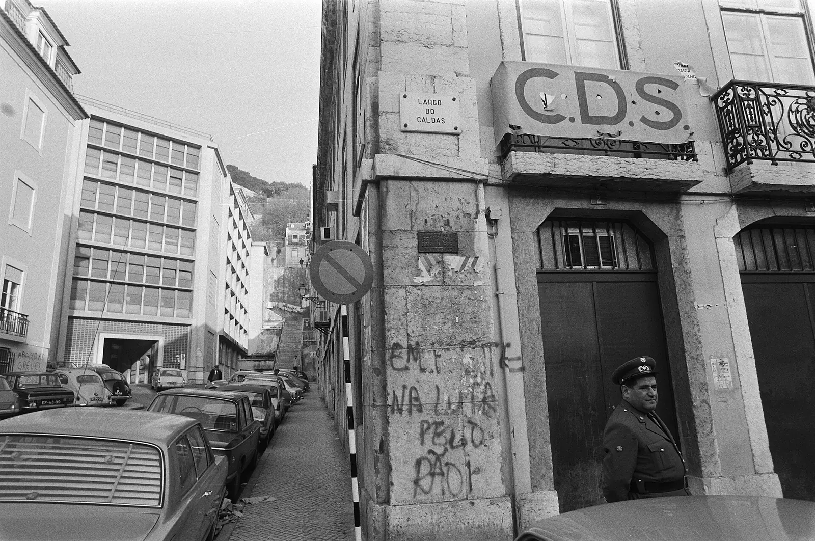 Fotografia a preto e branco de uma rua estreita com prédios antigos; à direita, uma fachada com as letras «CDS» e paredes cobertas de inscrições, e um homem à porta.