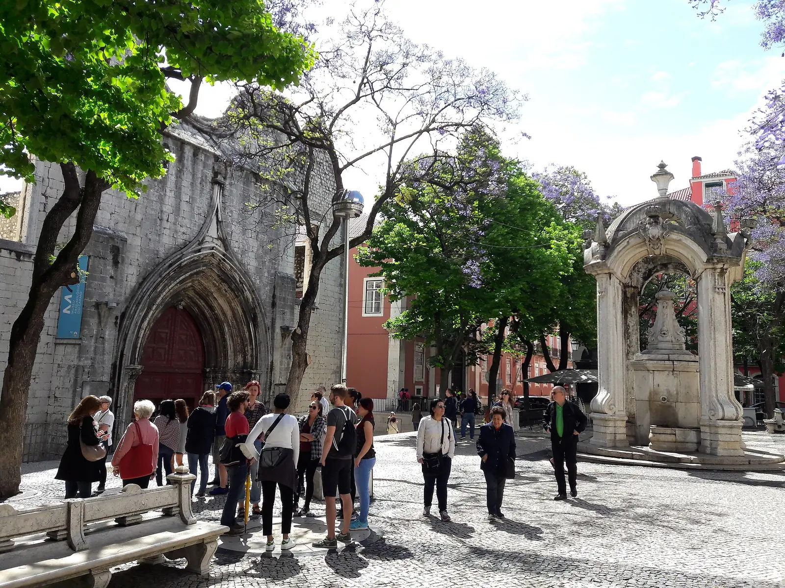 Largo arborizado com jacarandás em flor, o portal gótico de uma igreja em ruínas à esquerda, um chafariz de pedra à direita e grupos de pessoas a passear.