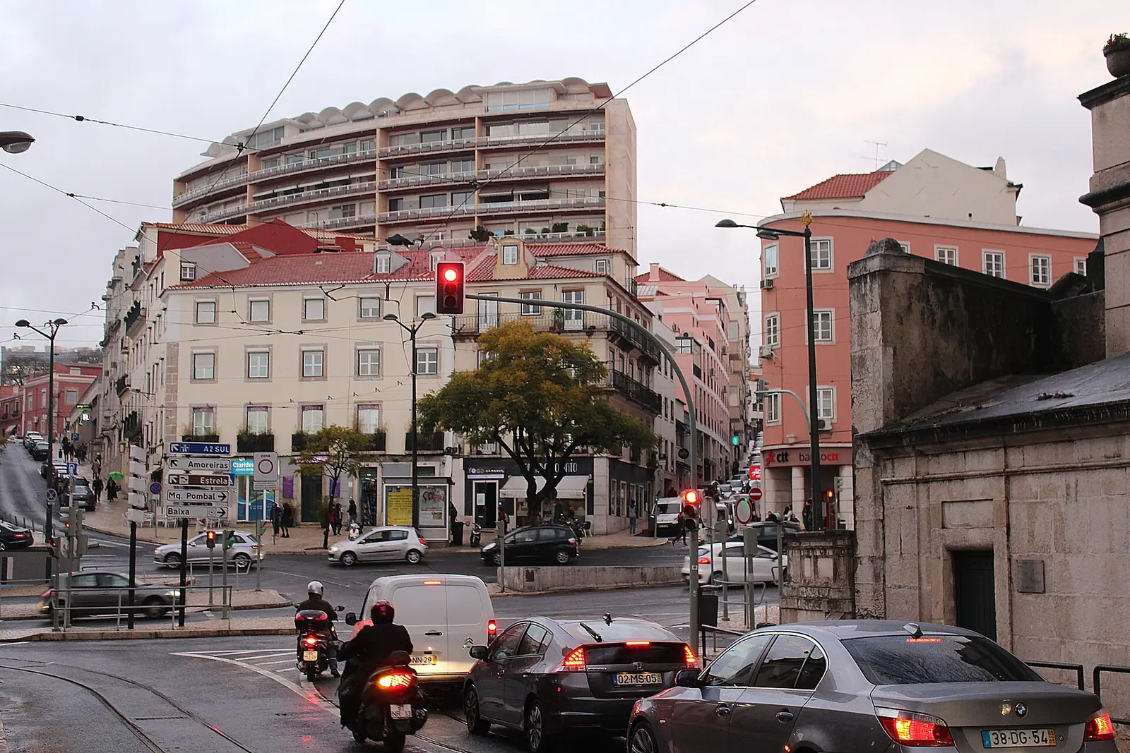 Cruzamento largo com carros e motas parados num semáforo, entre prédios de vários andares e cores, sob céu com nuvens.