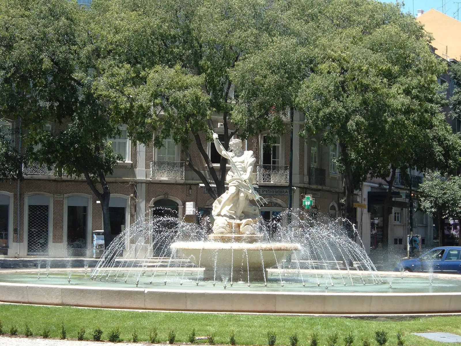 Chafariz redondo com uma escultura branca ao centro e repuxos de água, entre árvores frondosas e prédios de varandas.
