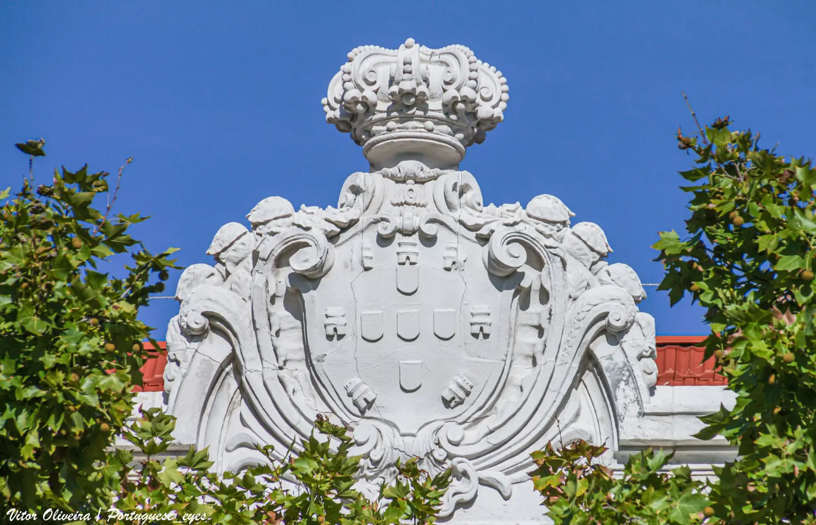 Escudo de pedra branca com coroa, esculpido em relevo, entre a folhagem verde de árvores e o céu azul.