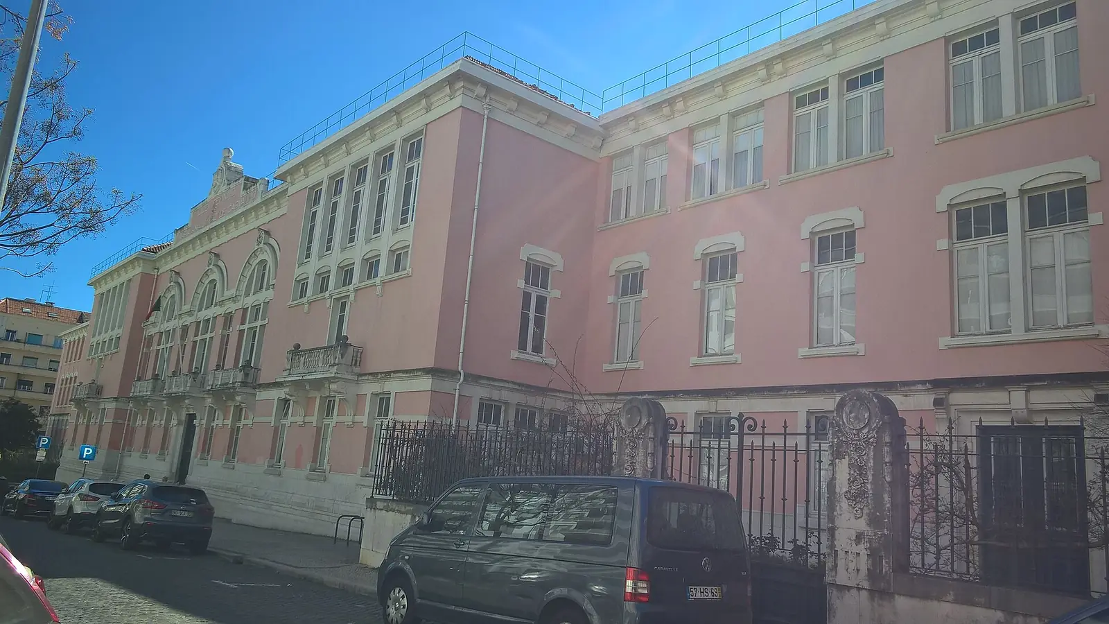 Edifício escolar comprido, cor-de-rosa, de dois andares, com janelas altas e um portão de ferro, numa rua com carros estacionados, sob céu azul.
