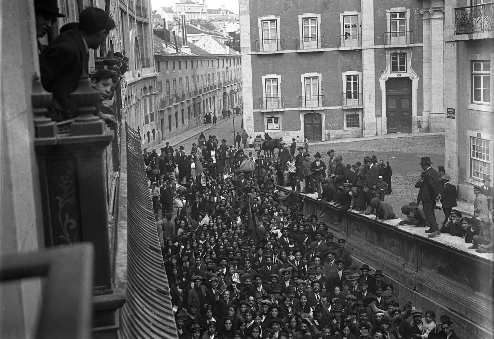 Fotografia a preto e branco tirada de uma varanda: a rua cheia de gente, de chapéu, diante de um prédio de muitas janelas.