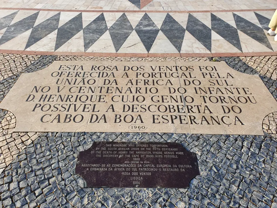 Laje de pedra clara com uma inscrição gravada em letras maiúsculas e a data de 1960, rodeada de calçada de pedras escuras; por cima, a orla da rosa-dos-ventos, em triângulos pretos e brancos; por baixo, uma placa escura de metal com o texto em inglês.