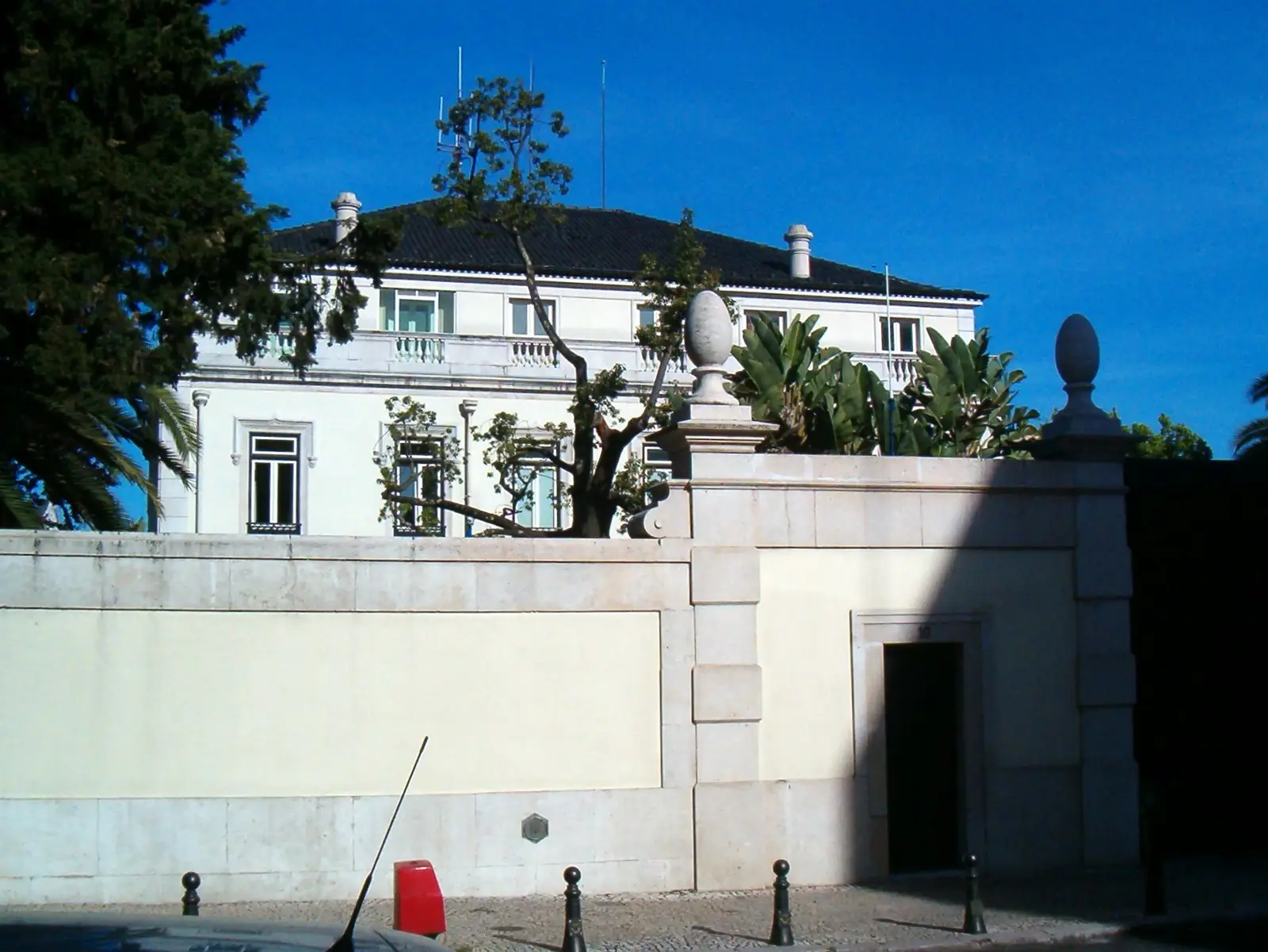 Fachada branca de um palacete com varandas e chaminés, entre árvores e palmeiras, por cima de um muro de pedra com uma porta escura.