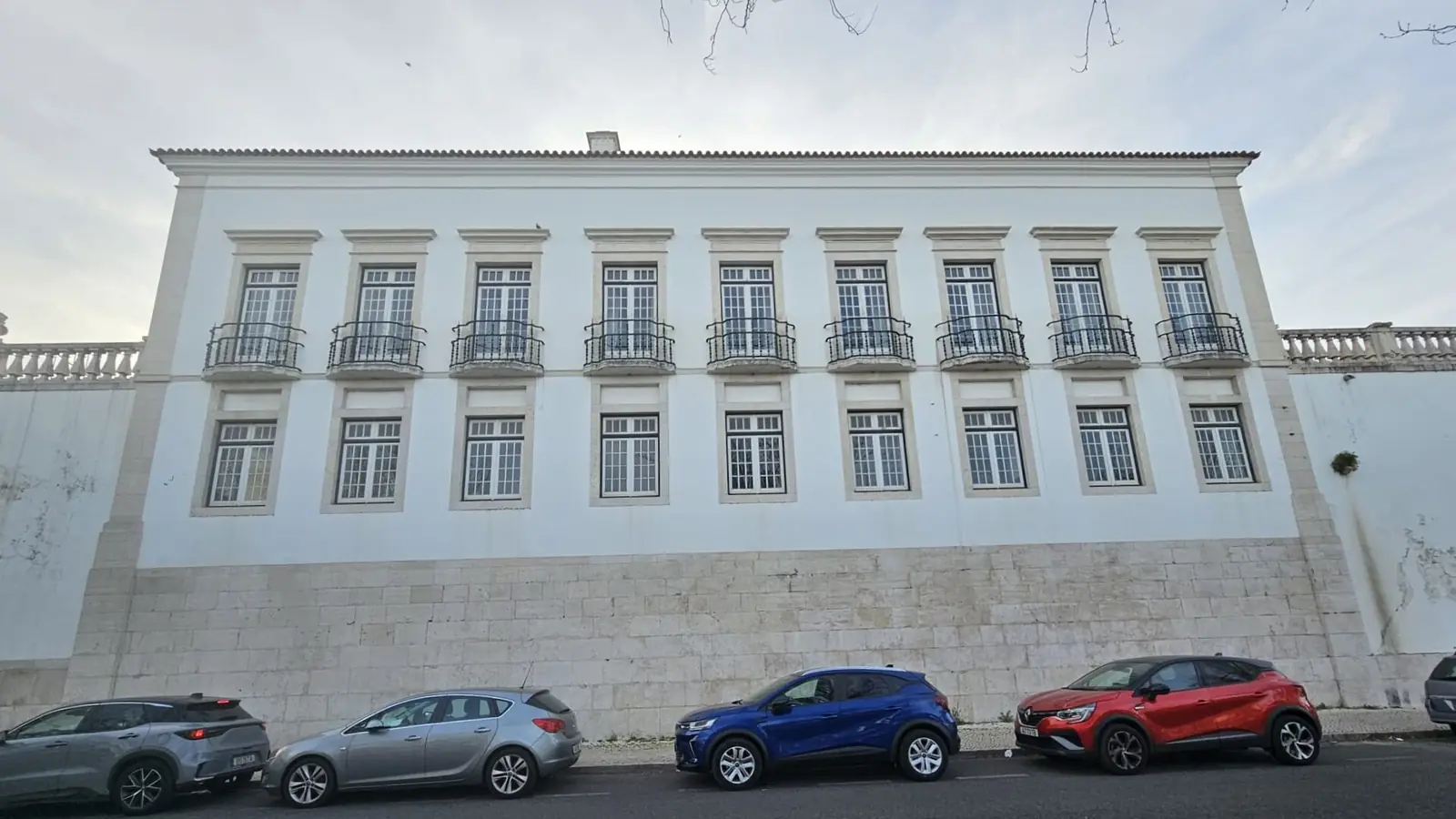 Fachada branca comprida de três andares com muitas janelas de moldura de pedra, sobre um embasamento de pedra, com carros estacionados à frente.