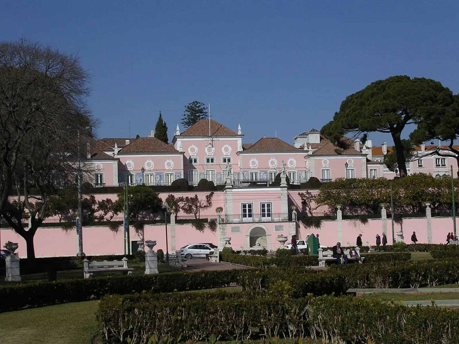 Fachada comprida cor-de-rosa, com janelas de moldura branca e telhados vermelhos, por trás de um muro e de um jardim de sebes baixas, com árvores e postes de iluminação.