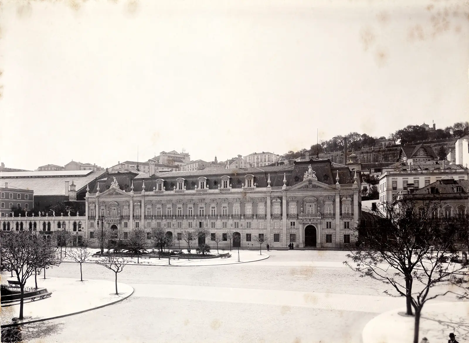 Fotografia antiga e desbotada da fachada comprida de um palácio, vista do outro lado de uma praça vazia, com árvores despidas à direita.