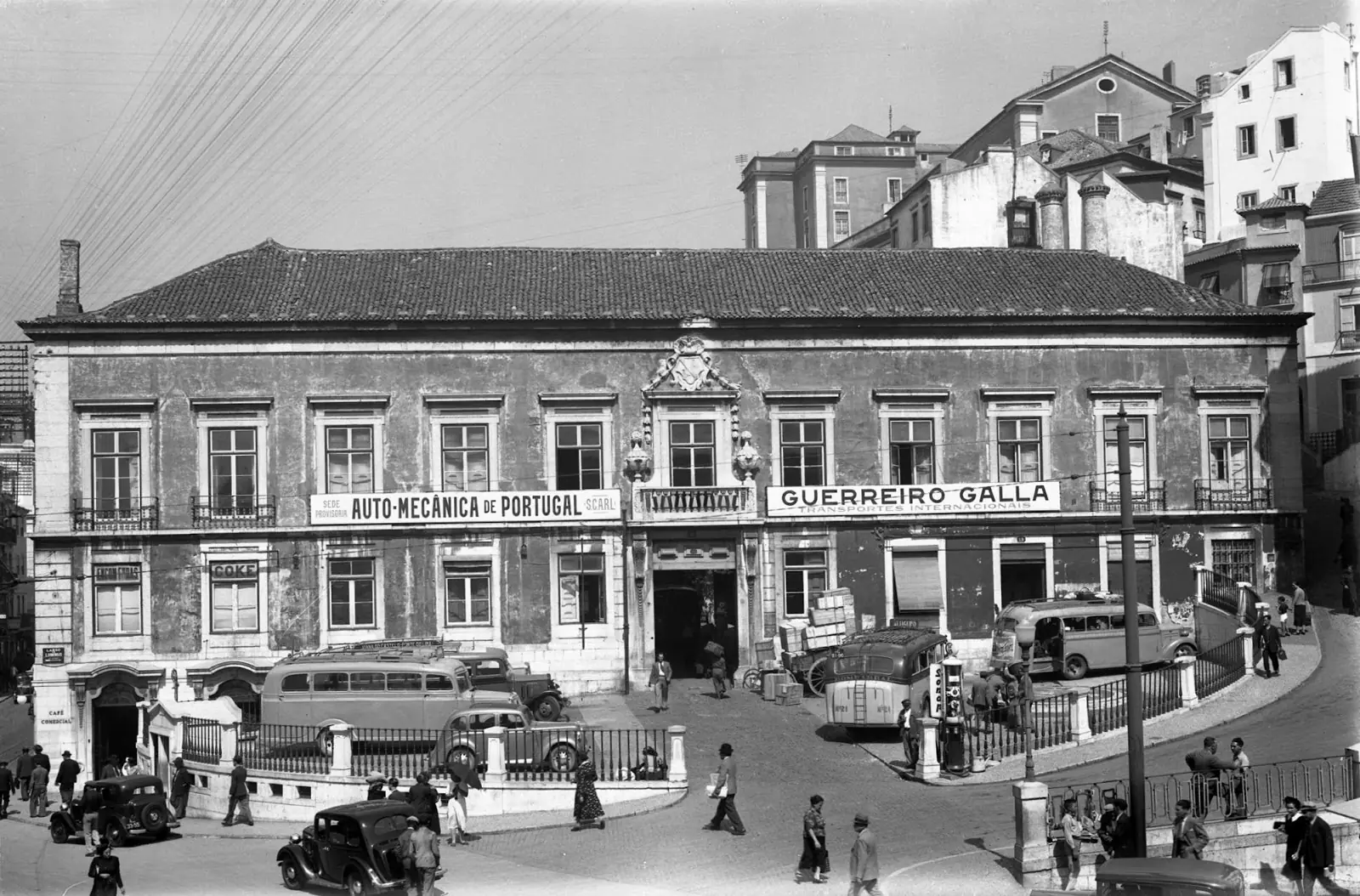 Fotografia a preto e branco da fachada comprida de um palácio de dois andares com um brasão ao centro, com autocarros e automóveis antigos estacionados à frente.