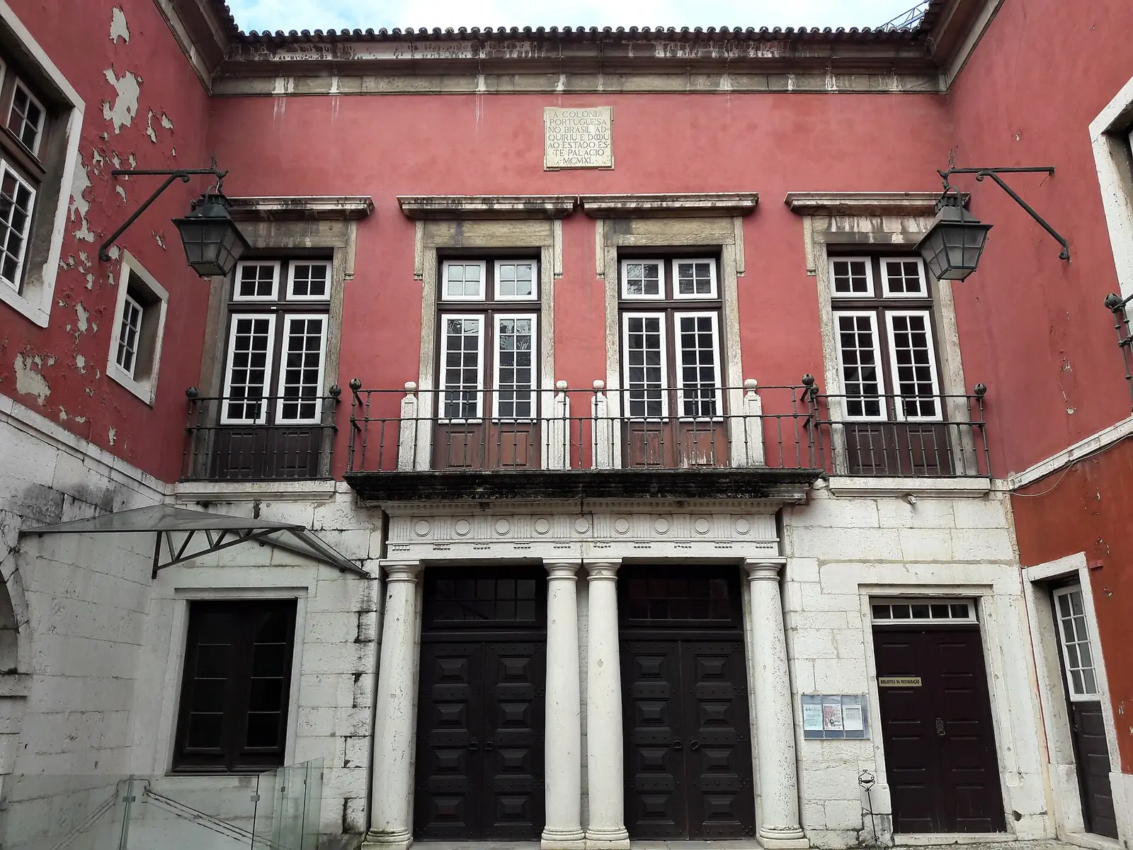 Pátio com fachadas vermelhas de dois andares, janelas e portas de moldura de pedra branca, uma varanda de ferro ao centro e candeeiros nas paredes.
