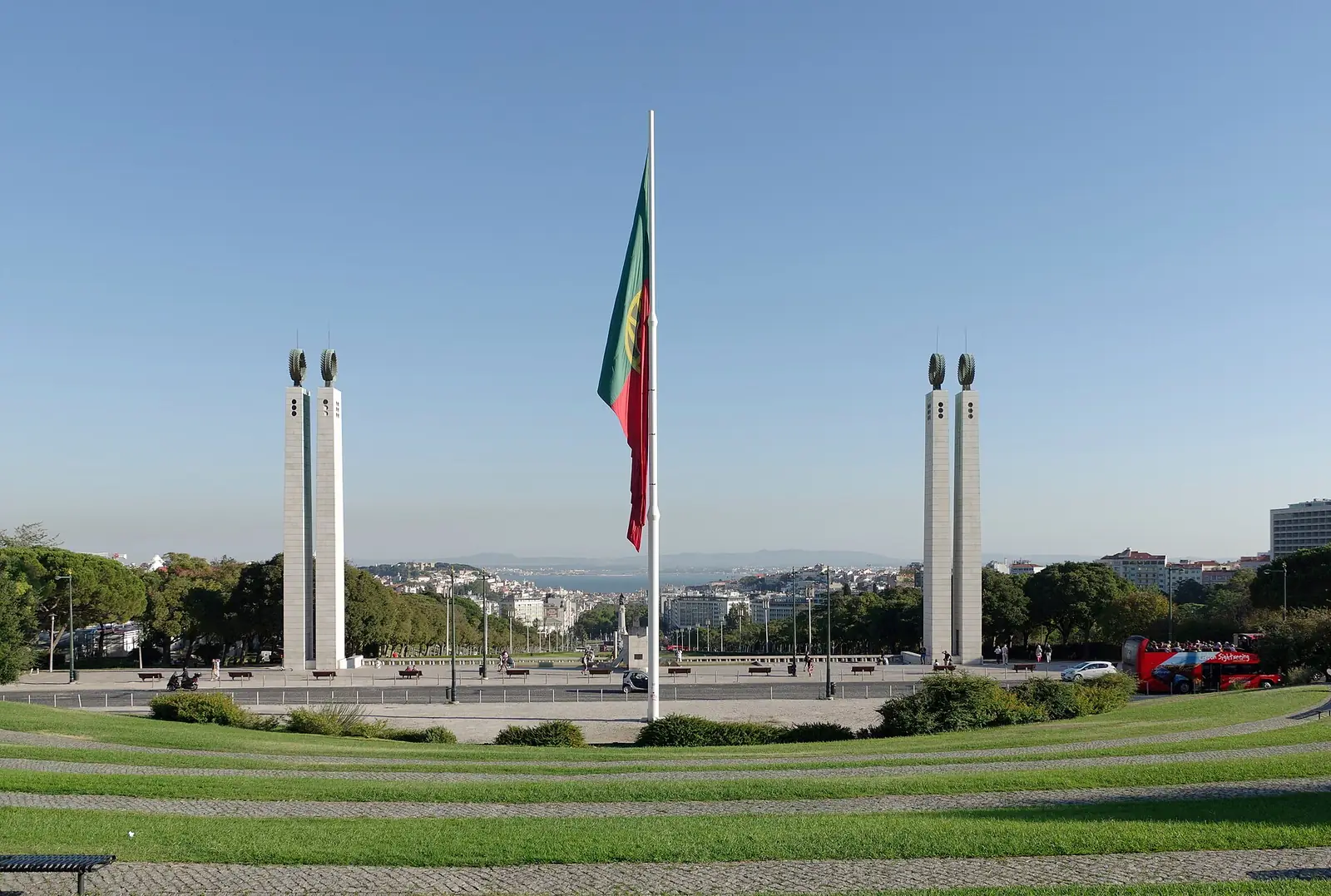 Bandeira portuguesa num mastro altíssimo entre dois pares de colunas brancas, num terraço de relva e calçada, com a cidade e o Tejo ao fundo.