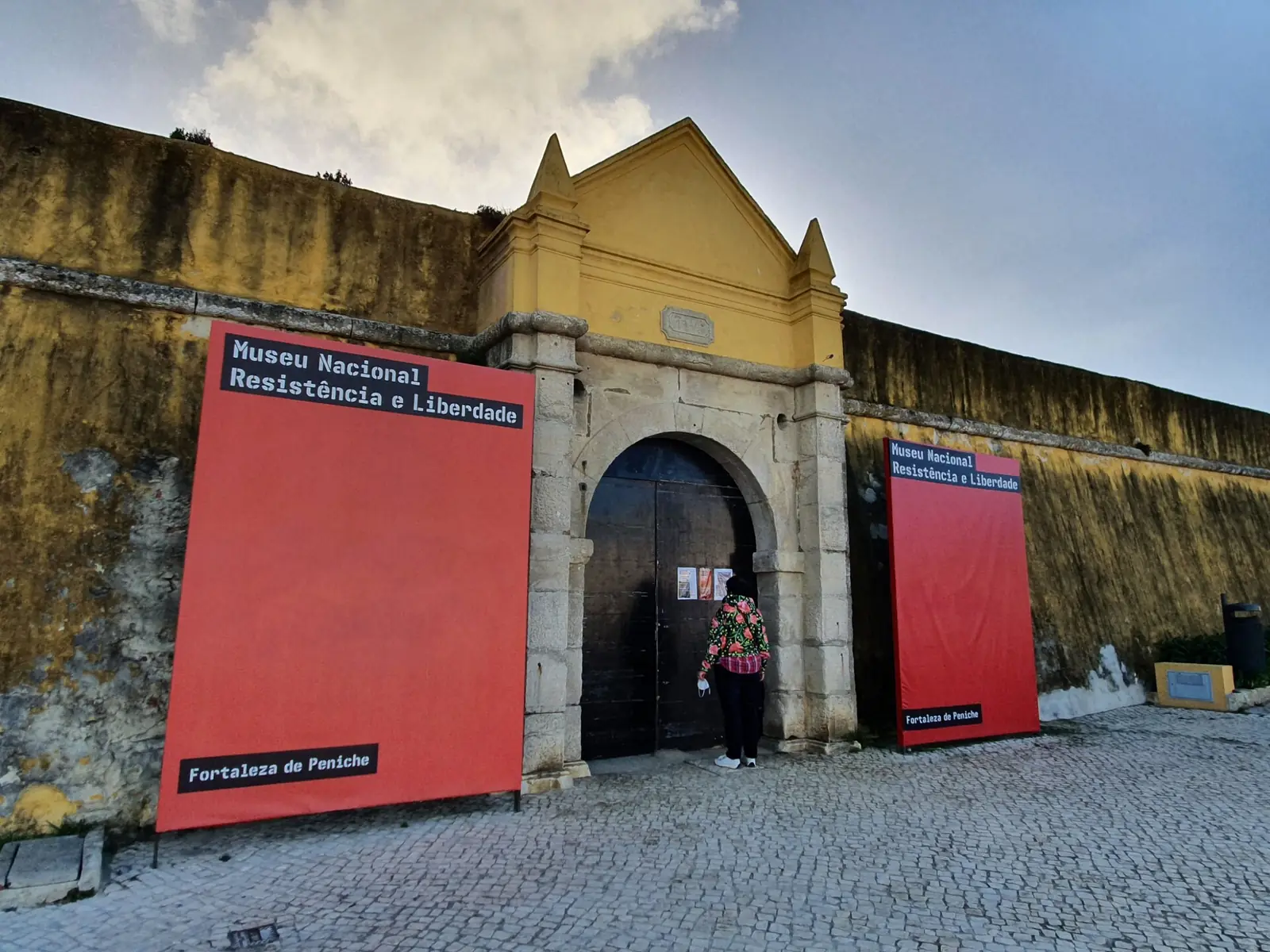 Portal de pedra com frontão amarelo e branco numa muralha antiga, com dois painéis vermelhos à entrada e um pequeno largo de calçada à frente.