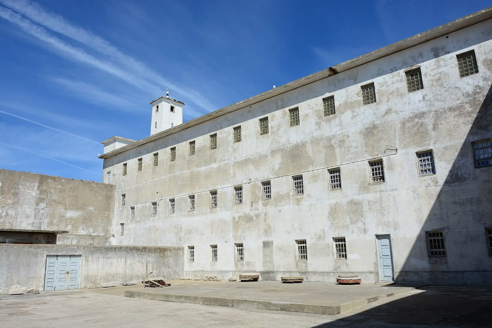 Edifício prisional comprido de três pisos, branco e cinzento, com fileiras de janelas pequenas e gradeadas, e uma torre de vigia ao fundo, sob céu azul.
