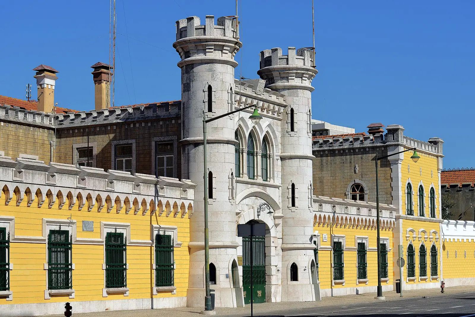 Edifício comprido amarelo com janelas gradeadas verdes e torres brancas de ameias junto à entrada, ao longo de uma rua com candeeiros.