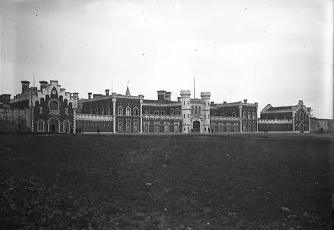 Fotografia antiga a preto e branco de um grande edifício comprido e acastelado ao fundo de um descampado, sob um céu claro.