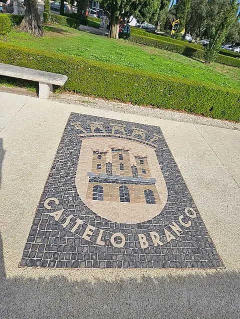 No chão claro de um passeio, um brasão em calçada portuguesa: um castelo amarelado de três torres, com uma coroa de torres por cima, sobre fundo de pedras pretas, e as palavras «Castelo Branco» em letras brancas; atrás, um banco de pedra, uma sebe e relva.