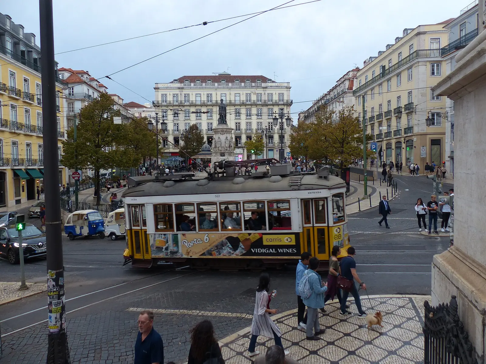 Praça larga de calçada clara com árvores e a estátua de Camões ao fundo, um elétrico amarelo a atravessar e gente a passear.
