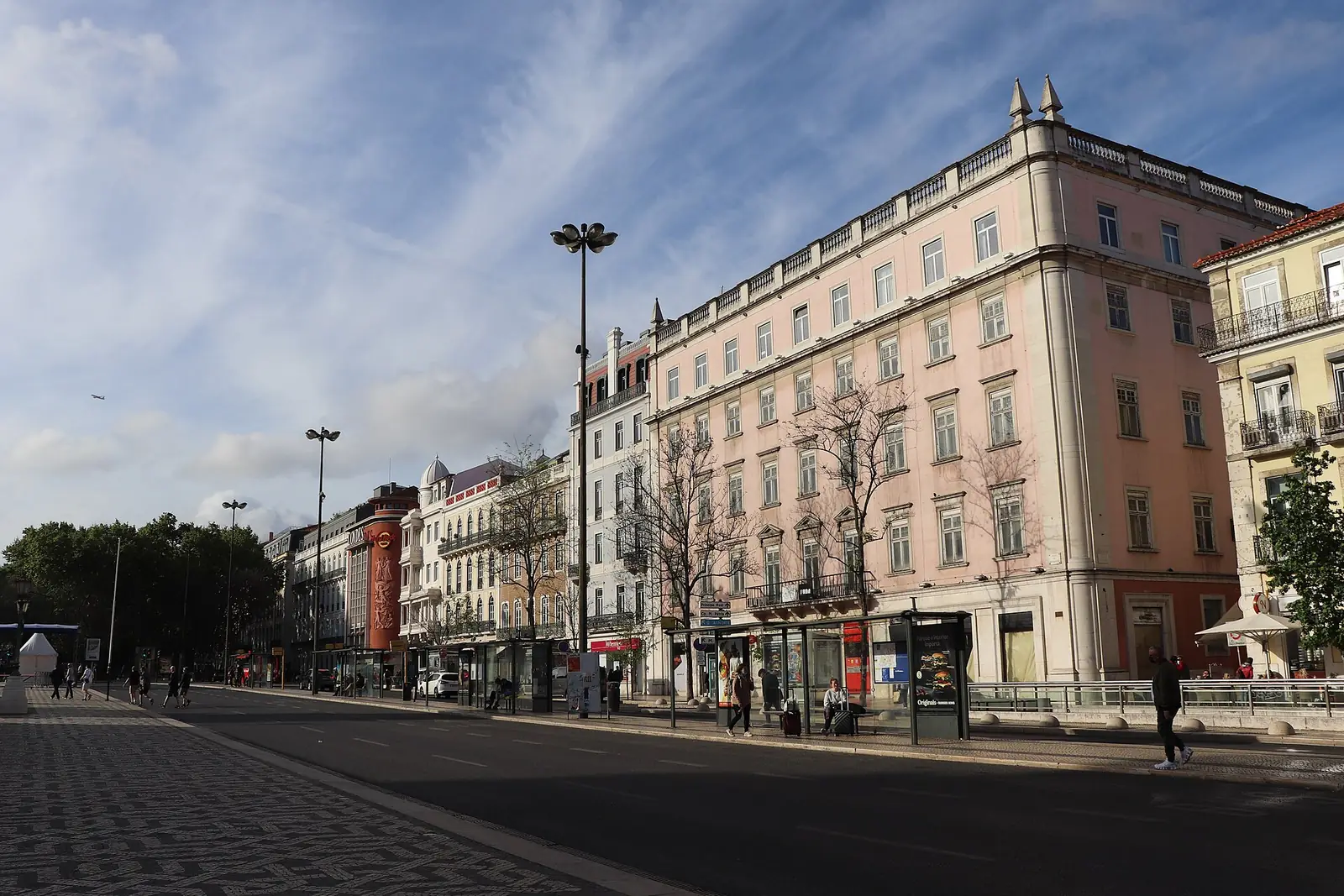 Fila de prédios altos de fachadas claras e rosadas ao longo de uma praça larga e quase vazia, com candeeiros altos e árvores, ao fim da tarde.