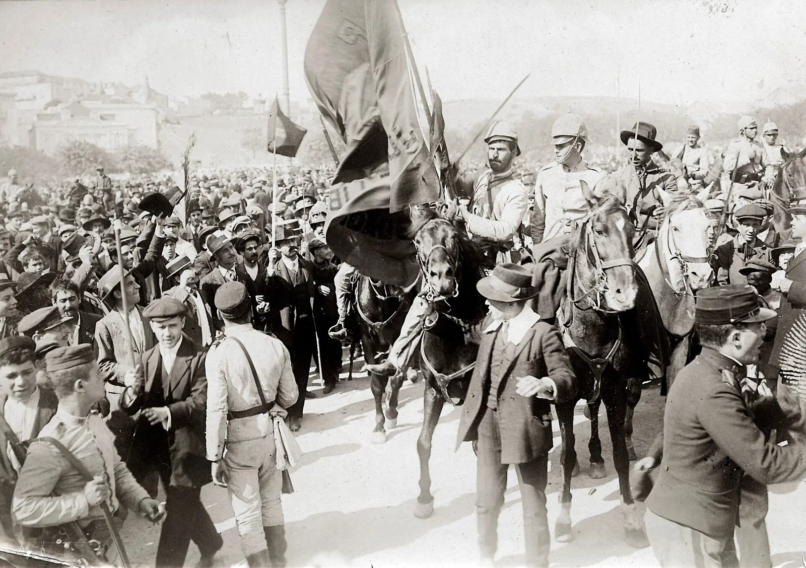 Fotografia antiga a preto e branco de uma multidão de homens de chapéu e de soldados a cavalo, com uma bandeira grande a esvoaçar por cima das cabeças.