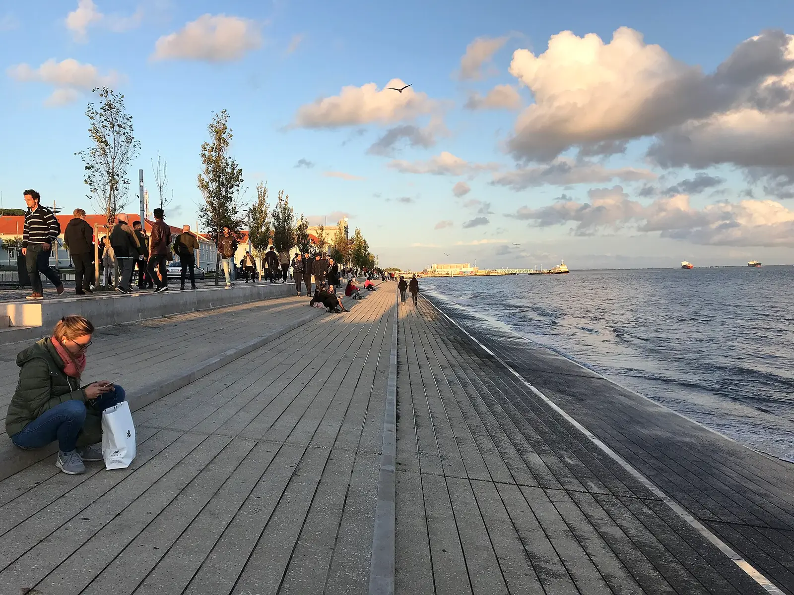 Passeio largo de madeira à beira-rio, com gente a caminhar e sentada; à direita, o Tejo; ao fundo, árvores e edifícios baixos sob nuvens.
