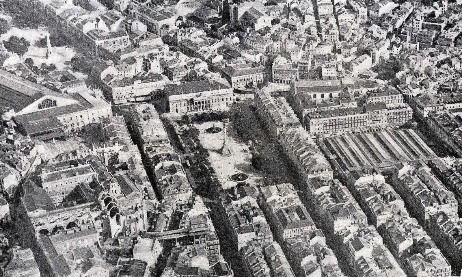 Fotografia aérea a preto e branco da Baixa: quarteirões regulares, uma praça comprida com a coluna ao centro e, ao lado, o grande mercado coberto de uma praça quadrada.