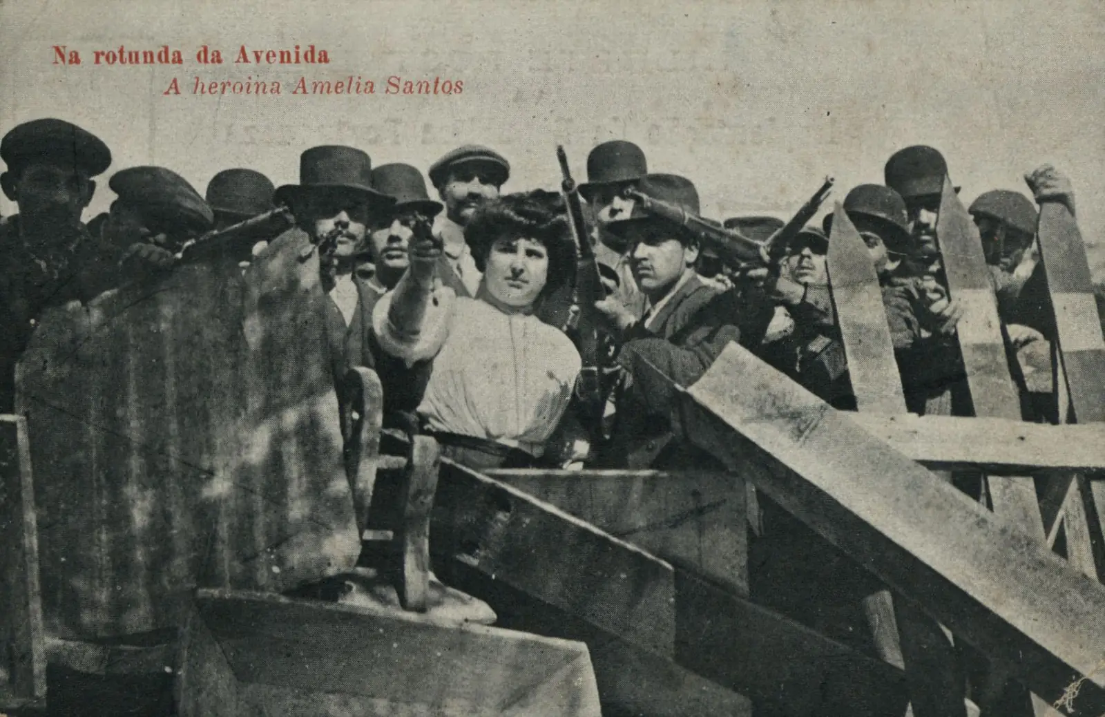 Postal antigo com o título impresso a vermelho: uma mulher jovem de cabelo apanhado, ao centro, entre homens armados de chapéu e boné, atrás de uma barricada de tábuas.