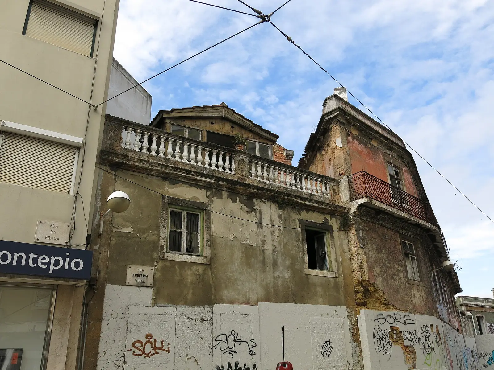 Prédio antigo de esquina, degradado e com inscrições nas paredes, com uma balaustrada no topo, visto de baixo, entre fios de elétrico e céu azul.