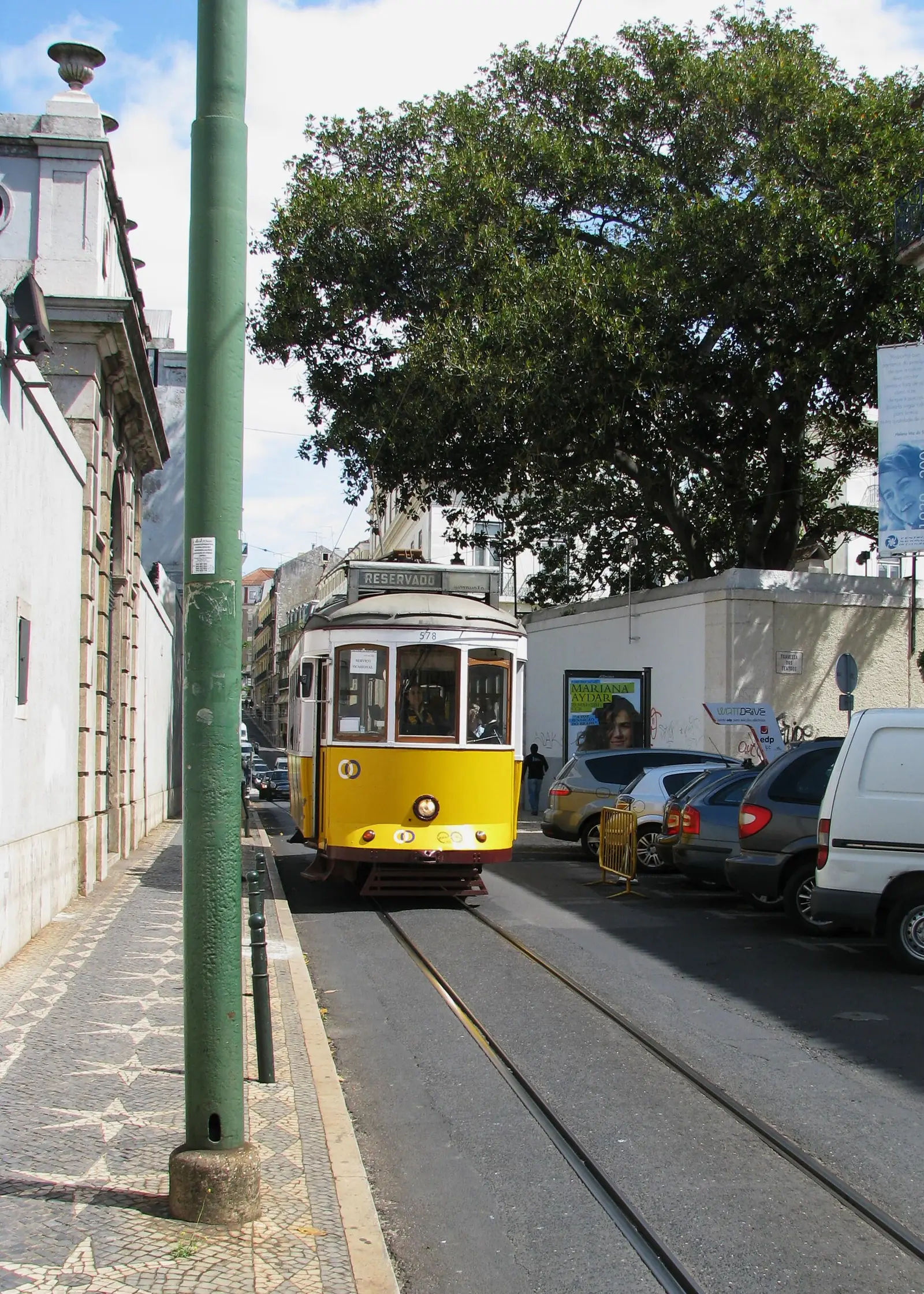 Elétrico amarelo numa rua com carris, entre fachadas claras à esquerda e carros estacionados à direita, debaixo de árvores.