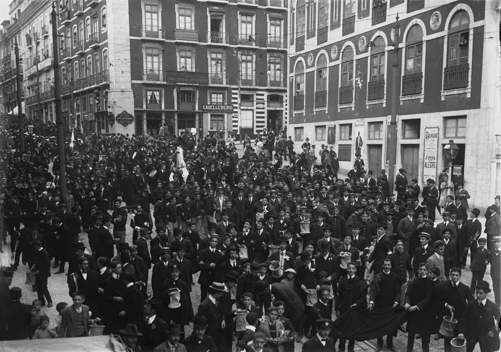 Fotografia a preto e branco de uma multidão de estudantes de capa e chapéu a encher a rua, diante de prédios de varandas.